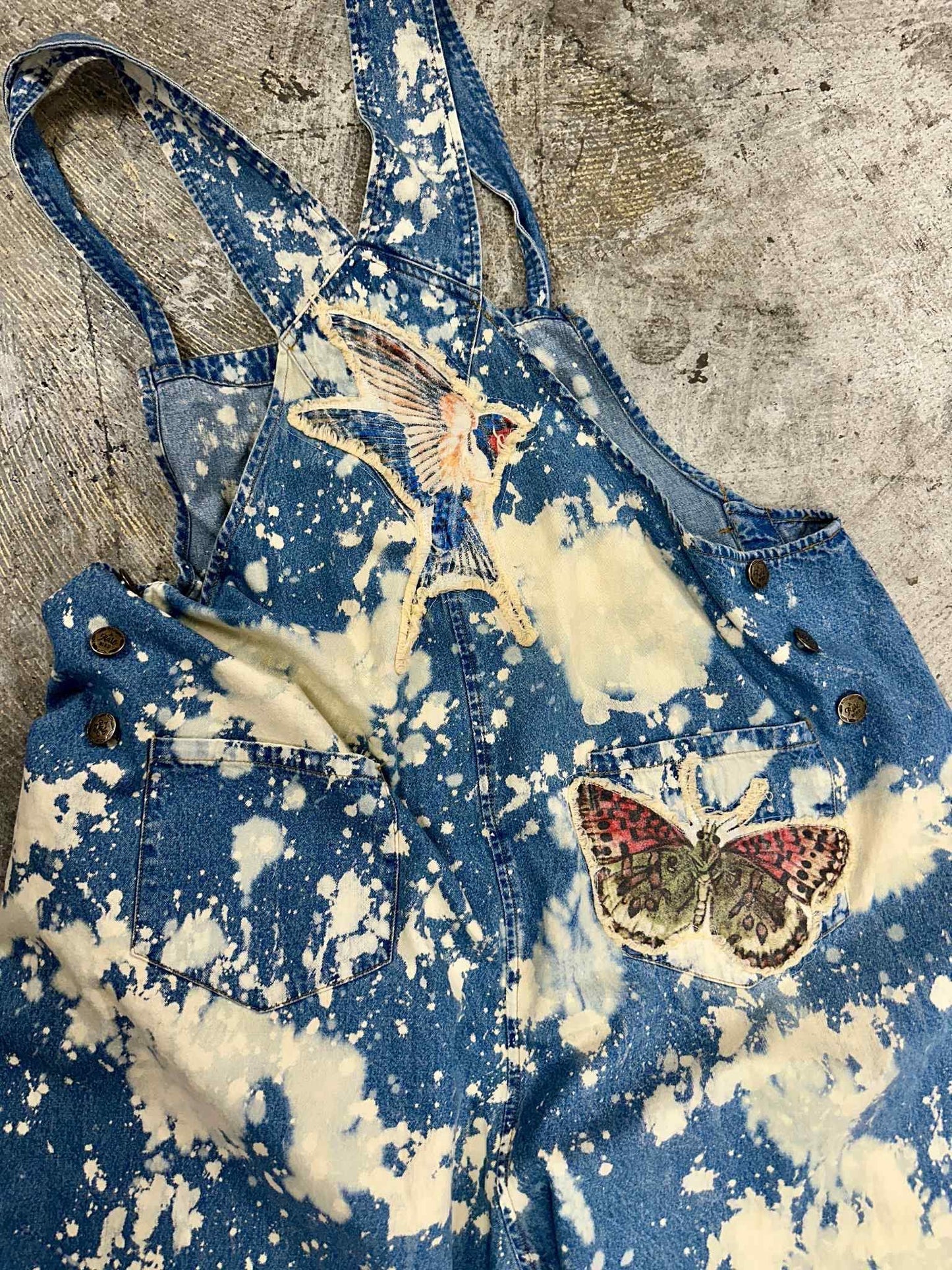 Blue denim overalls with white splatter design and butterfly patches on a textured surface. Boho Chic. Grunge Clothing. Unique Style. 
A Rare Bird Butterfly Overalls