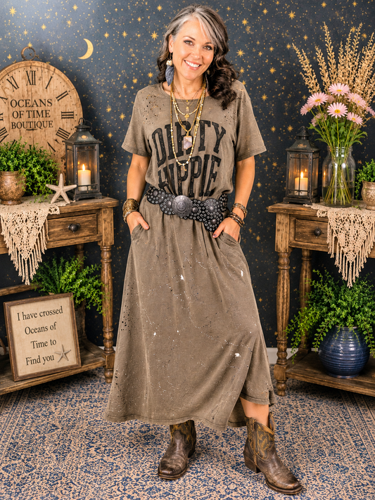 Woman wearing the Dirty Hippie Graphic Maxi Dress by Blue Velvet featuring distressed details, paint splatter accents, side pockets, and layered boho jewelry in a cozy boutique setting.
